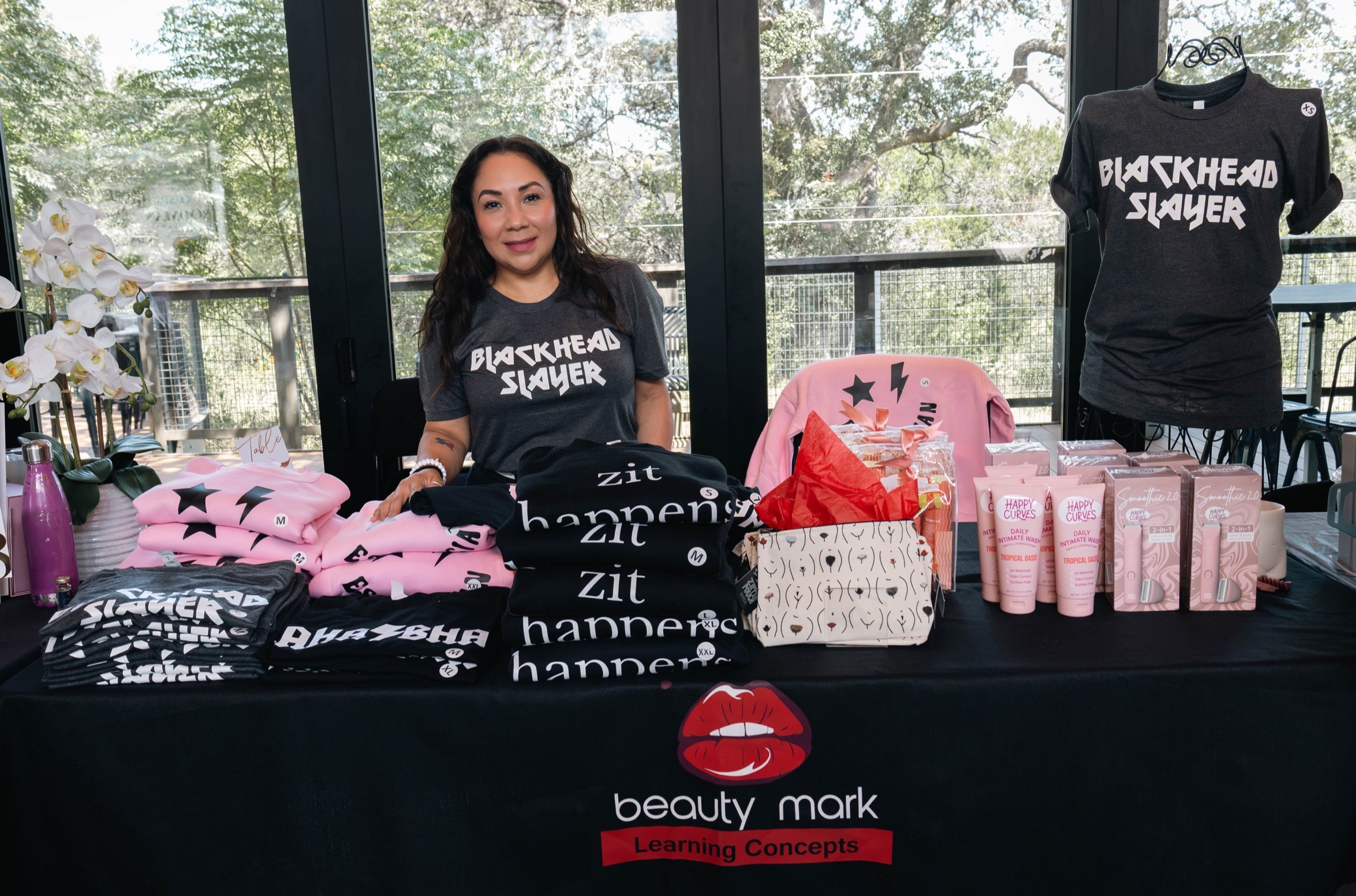 A smiling vendor stands behind a display of beauty merchandise and pink product packaging
